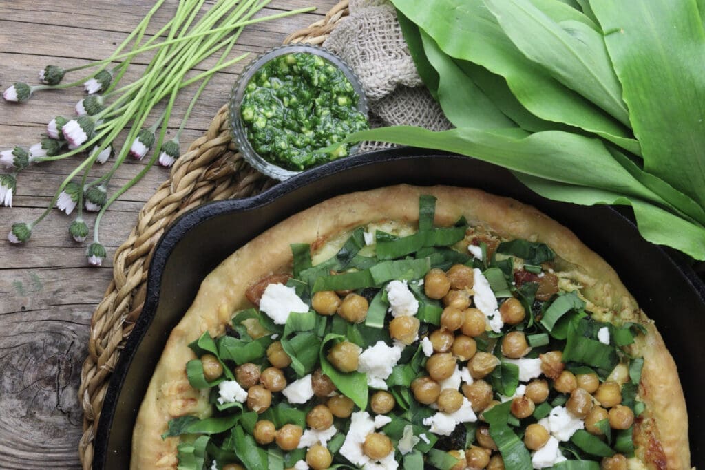 image of wild garlic pizza in a pan with roasted chickpeas and wild garlic leaves in the background